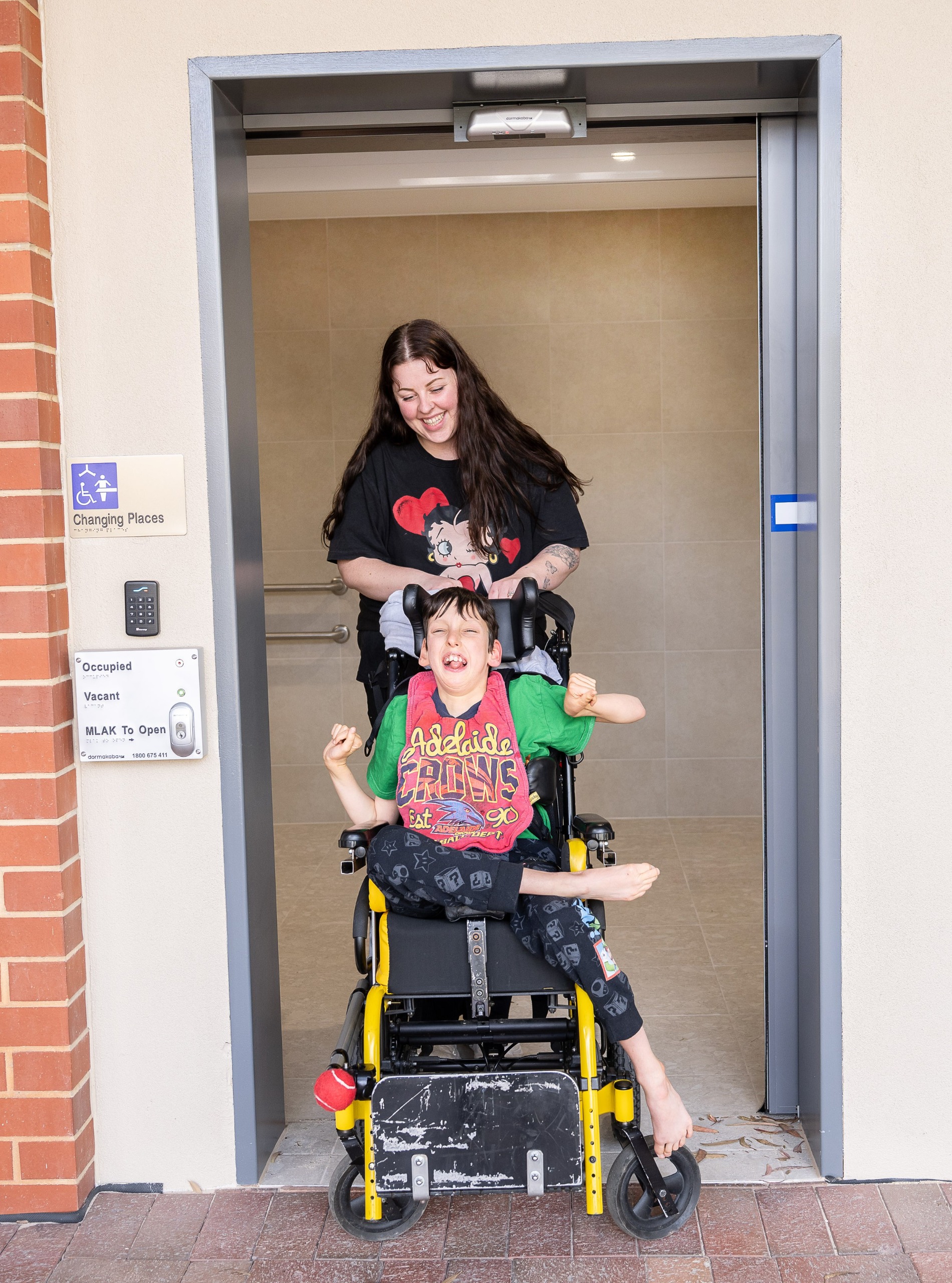 A woman smiling pushing a child in a wheelchair exiting a changing places toilet